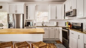 Eine moderne Küche mit weißen Schränken, einem Edelstahlkühlschrank, einer Mikrowelle und einem Backofen. An der Kochinsel stehen zwei Weidekorbstühle.