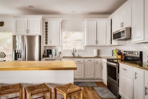 A modern kitchen with white cabinets, a stainless steel refrigerator, a microwave, and an oven. There are two wicker stools at the island.