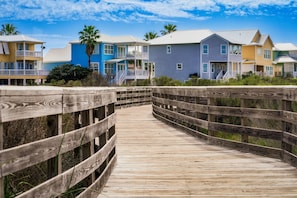 Une promenade en bois mène à des bâtiments colorés situés en bord de mer, dotés de balcons, sous un ciel dégagé.