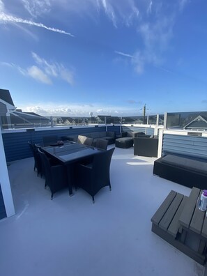 A rooftop patio with a glass balustrade, a dining table with chairs, and a wooden bench.