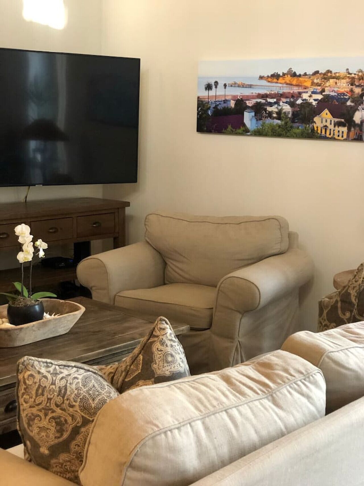 A living room with a beige sofa, patterned cushions, a wooden coffee table with a potted orchid, a flat-screen TV, and a coastal landscape painting on the wall.