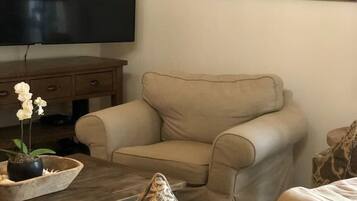 A living room with a beige sofa, patterned cushions, a wooden coffee table with a potted orchid, a flat-screen TV, and a coastal landscape painting on the wall.