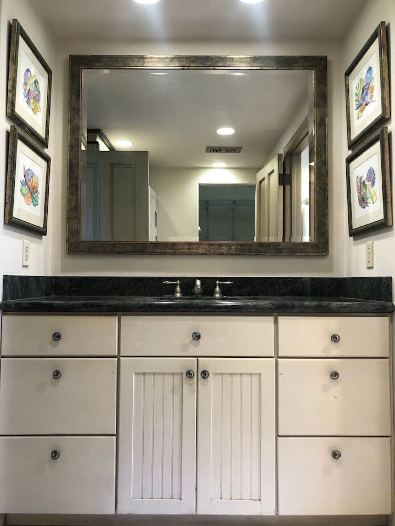 A bathroom with a large mirror, white cabinets, and framed artwork on the walls.
