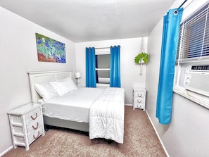 A bedroom with a bed, a nightstand, a dresser, a window with blinds, a painting on the wall, and a potted plant.