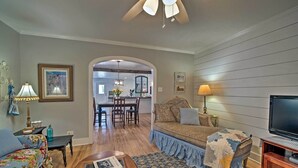 A living room with a sofa, coffee table, and TV, wooden flooring, and a kitchen in the background. - Pet-Friendly Brenham Cottage w/ Yard! (Brenham)