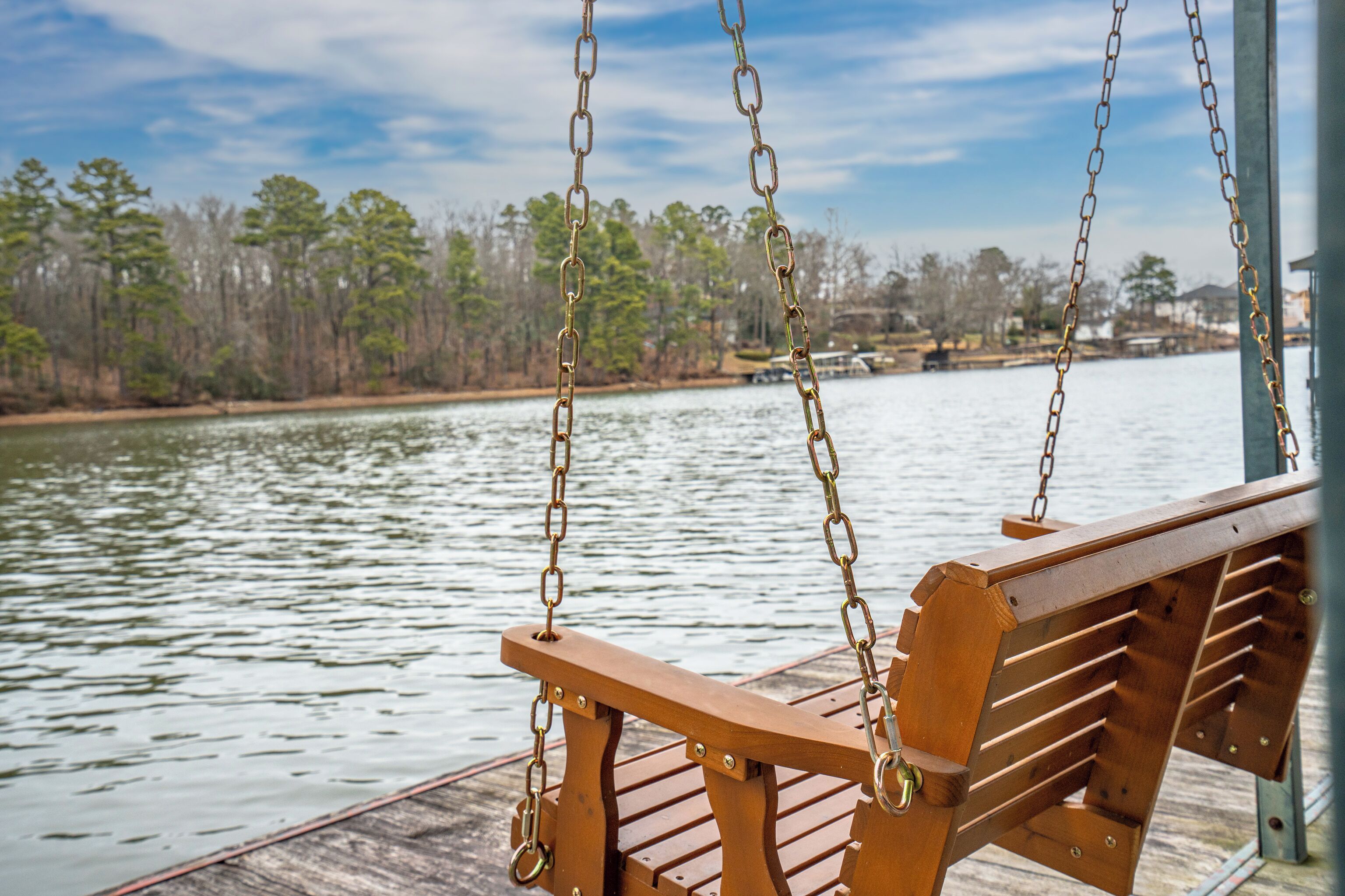 A wooden swing chair on a dock with a view of a calm lake and trees in the background.