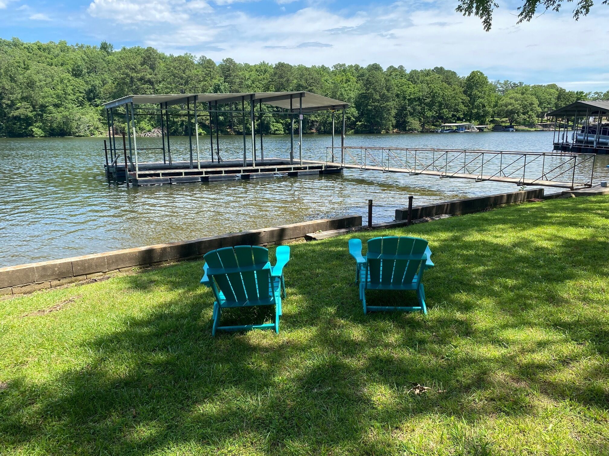A lakeside deck with two Adirondack chairs facing the water, surrounded by a grassy area and a dock extending into the lake.