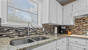 Cocina con armarios blancos, encimera de granito, pileta doble y ventana con persianas.