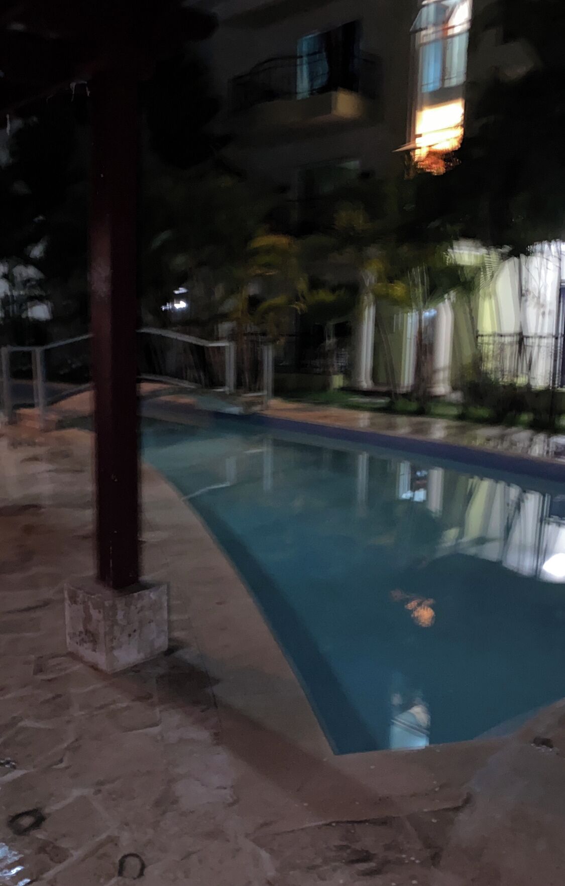 A swimming pool with a stone deck, surrounded by greenery and a building with balconies.