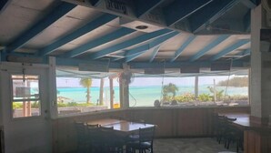 A restaurant with a view of the ocean, featuring a high ceiling with blue beams, tiled floor, and several tables and chairs.