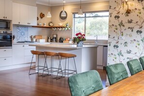 A modern kitchen with white cabinets, a central island with stools, a dining table with green chairs, and a window with a view of trees. - The Farmstead - Family Accommodation  (Dixons Creek)