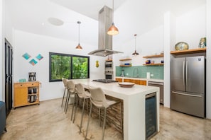 A modern kitchen with a central island, bar stools, stainless steel appliances, and a large window with a view of greenery.