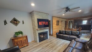 Un salon doté d’une cheminée en pierre, de canapés en cuir, d’un meuble en bois et d’un téléviseur fixé au mur, sur lequel est affiché le logo « NETFLIX ».