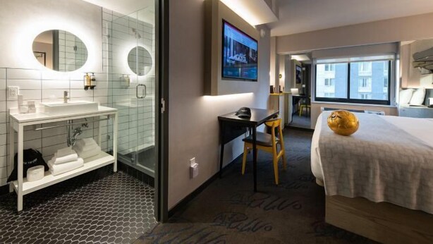 A hotel room with a glass shower enclosure, a white sink, a flat-screen TV mounted on the wall, a bed with a white bedspread, and a large window.