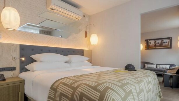 A hotel room with a bed, bedside table, lamp, and a wall-mounted air conditioner. - Pestana CR7 Times Square (New York)