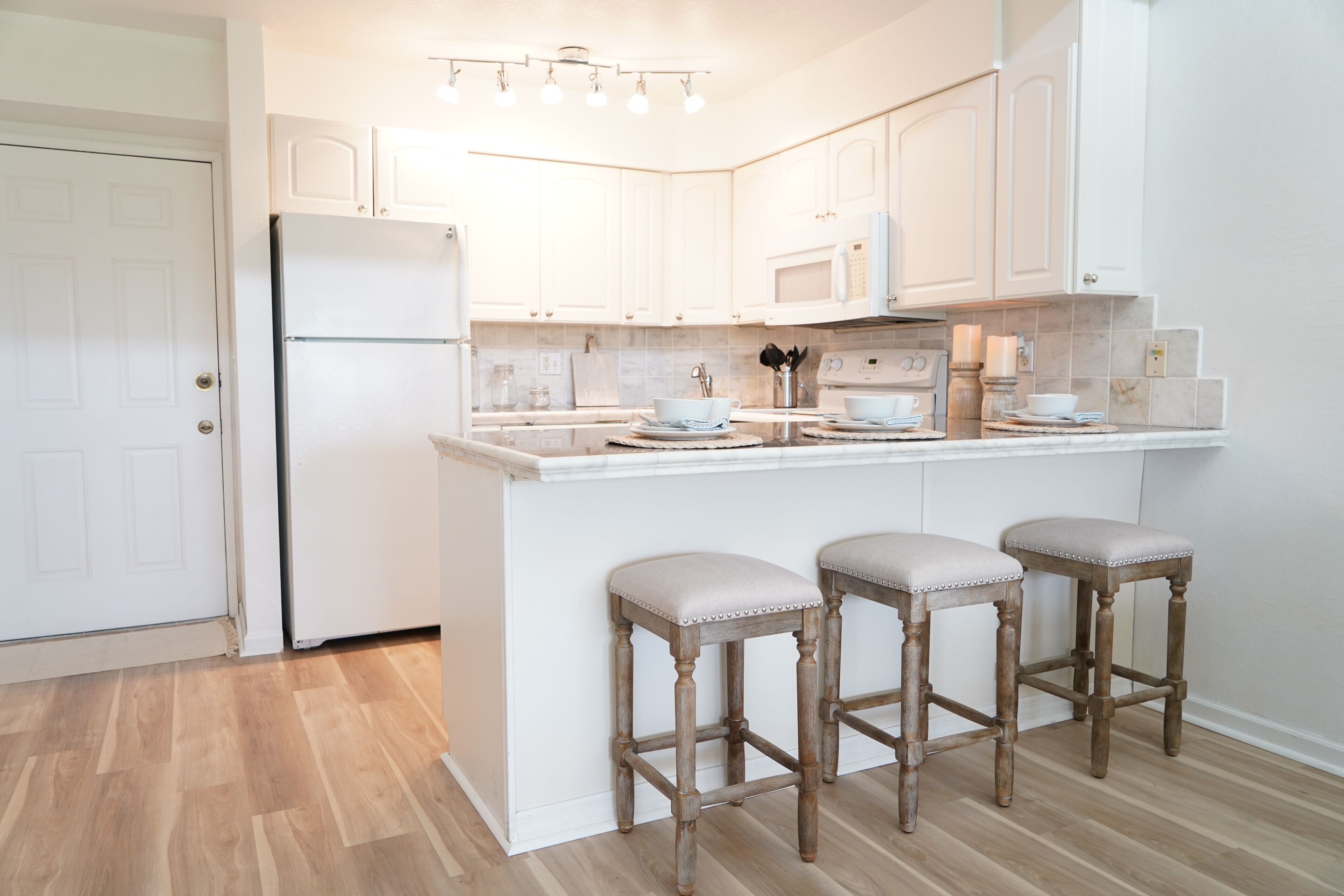 A kitchen with white cabinetry, a built-in microwave, a refrigerator, and three bar stools with cushioned seats.