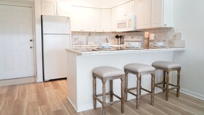 A kitchen with white cabinetry, a built-in microwave, a refrigerator, and three bar stools with cushioned seats. - 🏖️Just Remodeled! The Ocean Cottage at Ocean Village Beach Condo (Fort Pierce)