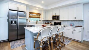 A modern kitchen with white cabinetry, stainless steel appliances, a central island with bar stools, and a herringbone-patterned backsplash. - Sanibel Surfsound Home - Heated pool and easy walk to the beach (Sanibel)