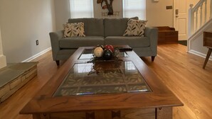 A living room with a glass-top coffee table, a gray sofa with patterned cushions, and a decorative tree wall art piece. - Beautiful House 4 ENTIRE Family near Suntrust Park (Marietta)