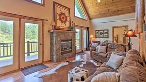 A spacious living room with a stone fireplace, wooden ceiling, and large windows overlooking a deck. - Idyllic Hill City Home - Hike, Bike & ATV! (Hill City)