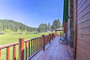 A wooden deck with a view of a green field and trees, a stone wall, and a rocking chair. - Idyllic Hill City Home - Hike, Bike & ATV! (Hill City)