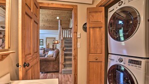 A laundry room with a front-loading washer and dryer, wooden doors, and a staircase leading to a living area with a couch and a lamp. - Idyllic Hill City Home - Hike, Bike & ATV! (Hill City)