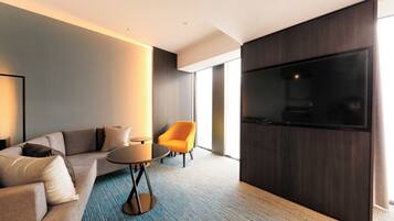 Una habitación de hotel moderna con un sofá, una mesita redonda pequeña, una silla naranja brillante y una pantalla de televisión grande.