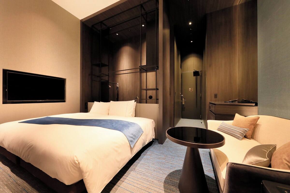 A modern hotel room with a large bed, a smaller sofa, a TV mounted on the wall, and a separate bathroom area.