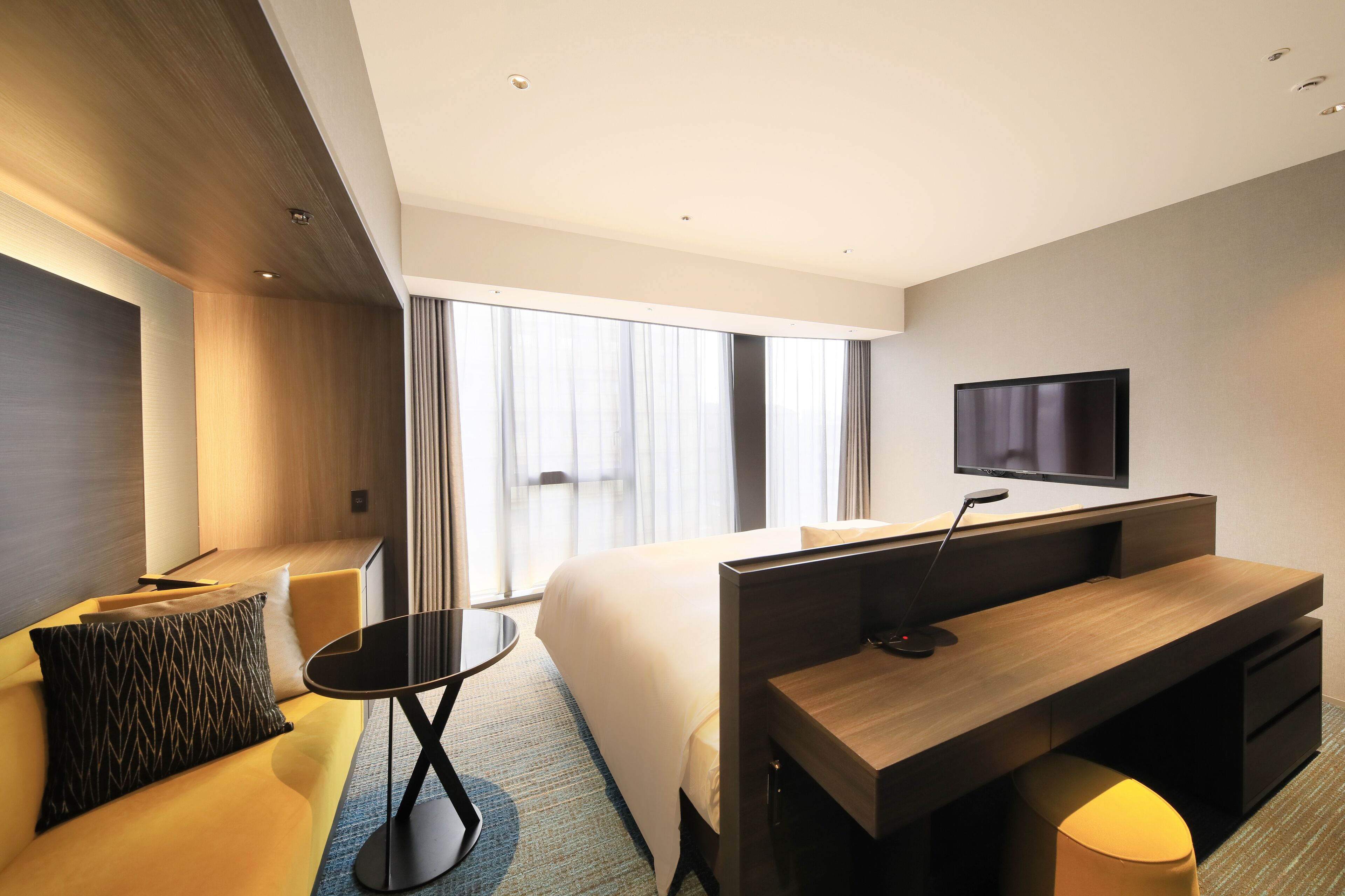 A modern hotel room with a large bed, a desk, a television, and a seating area with a sofa and a small table.
