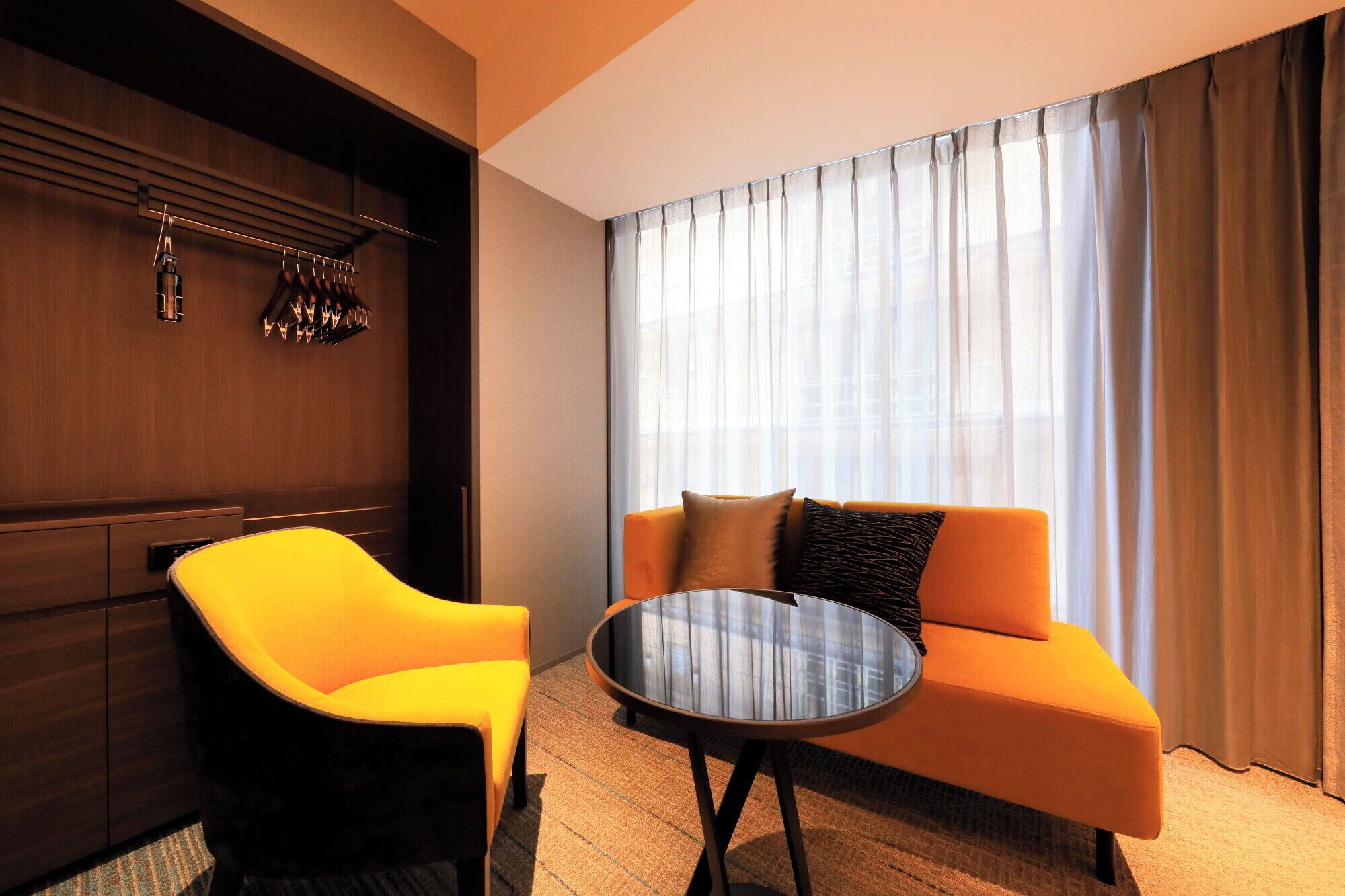 A modern hotel room with a sofa, a chair, a round table, and a large window with sheer curtains.