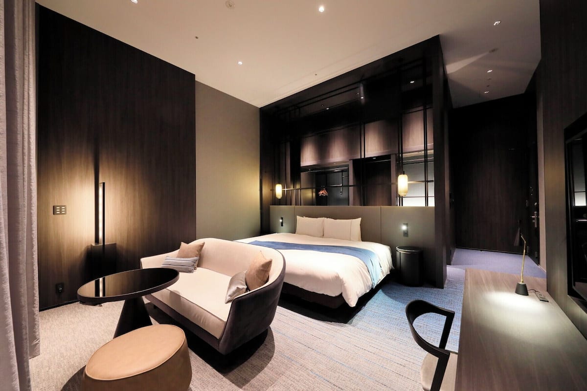 A modern hotel room with a large bed, a sofa, a small round table, and a desk.