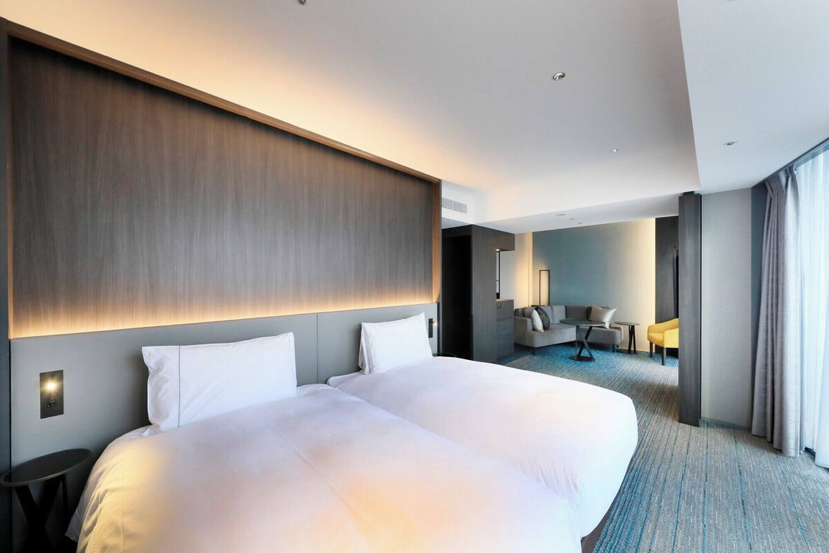 A modern hotel room with a large bed, a seating area with a sofa and armchairs, and a view of the city.