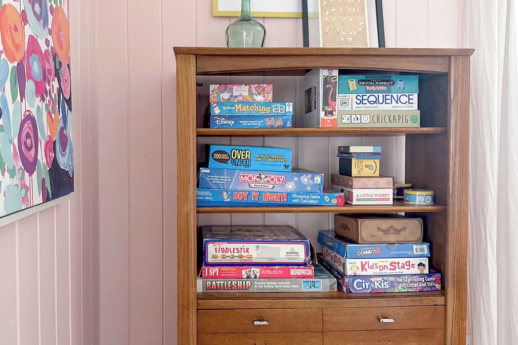 Uma estante de madeira com vários jogos de tabuleiro, incluindo 'Monopoly' e 'Battleship'.