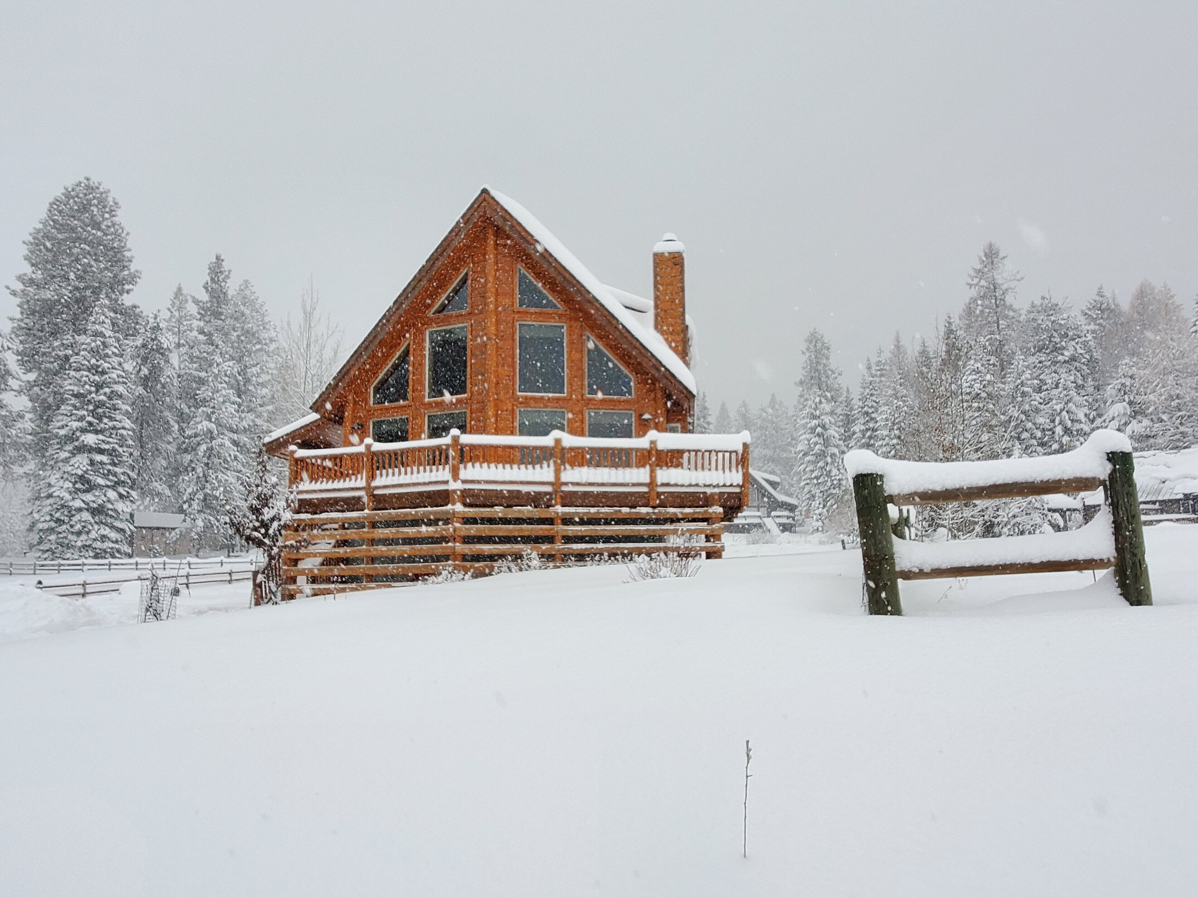 A log cabin with a wooden deck and a chimney, surrounded by snow-covered trees.