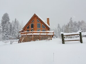 A log cabin with a wooden deck and a chimney, surrounded by snow-covered trees. - Gorgeous Lake view log cabin near Tamarack ski resort (Donnelly)