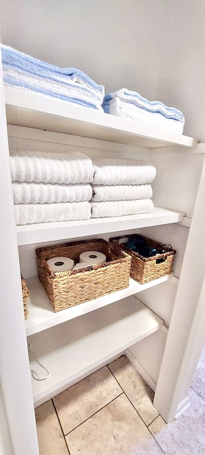 A white storage unit with shelves holding neatly folded white towels and blue towels, and wicker baskets containing items.