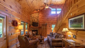 A cozy cabin interior with a fireplace, wooden walls, a ceiling fan, and a large window. - Amen Corner - Close to Boone! Hot Tub, Pet Friendly, Ping Pong, Foosball, Mountain Views (Vilas)