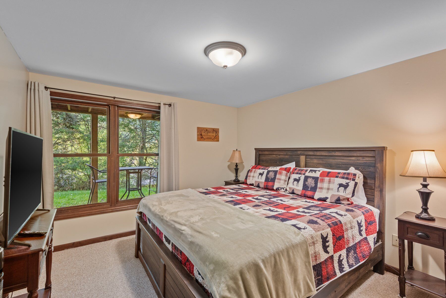 A bedroom with a large bed, a TV, a window with a view of greenery, and a ceiling light.