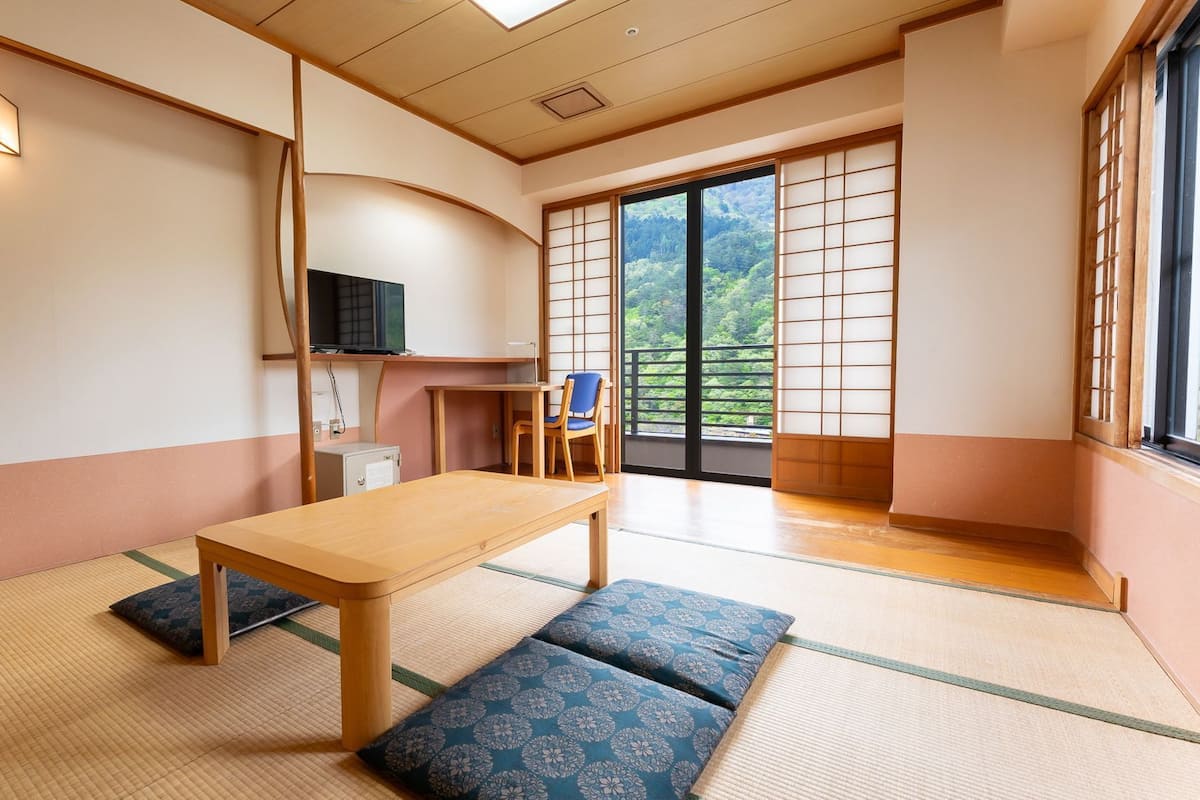 Une pièce de style japonais traditionnel, avec un sol recouvert de tatamis, une table basse, une chaise et une porte coulissante donnant sur un balcon d’où l’on peut admirer la verdure.