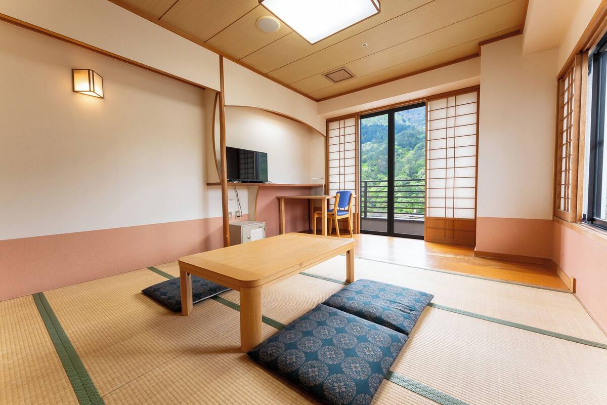 Une pièce de style japonais traditionnel, avec un sol recouvert de tatamis, une table basse, des coussins, un téléviseur et des portes coulissantes donnant sur un balcon avec vue sur la verdure.