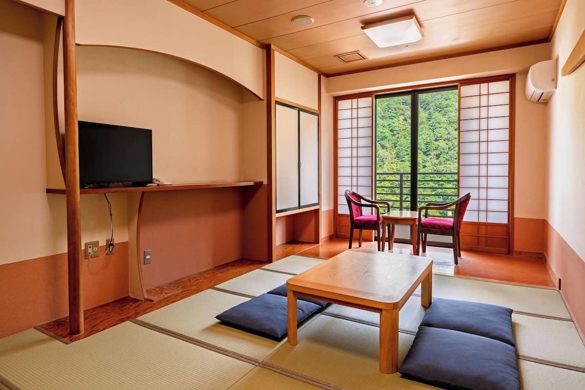 Une pièce de style japonais traditionnel, avec un sol recouvert de tatamis, une basse table en bois, deux chaises, un téléviseur posé sur une étagère et une porte coulissante donnant sur un paysage verdoyant.