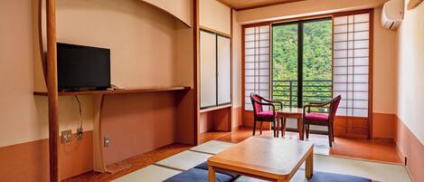 A traditional Japanese room with tatami flooring, a low wooden table, two chairs, a television on a shelf, and a sliding door with a view of greenery.