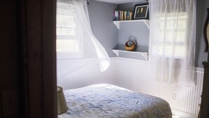 Een slaapkamer met een bed, een raam met dunne gordijnen, een plank met boeken en een mand, en een radiator.