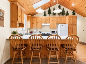 Una cocina con armarios de madera, encimera blanca, estufa, microondas y tres sillas de madera.