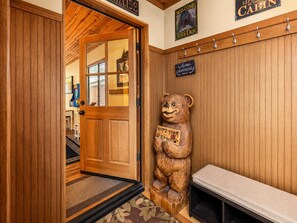 A wooden cabin interior with a bear statue holding a sign, a bench, and a door leading to a room with a window.