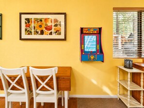 A room with a yellow wall, a framed floral artwork, a Pac-Man arcade machine, a wooden desk, two white chairs, and a window with blinds. - Moonridge Heaven - Close to Slopes, Fireplace, Deck, Barbecue (Big Bear Lake)