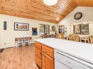A kitchen with wooden cabinets, a dishwasher, a dining area with chairs, a clock on the wall, and a window with blinds. - Moonridge Heaven - Close to Slopes, Fireplace, Deck, Barbecue (Big Bear Lake)