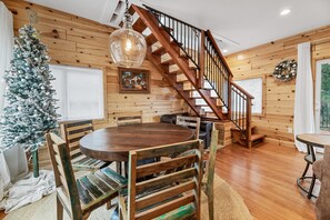 A cozy room with a wooden staircase, a dining table with chairs, a Christmas tree, and a large window.