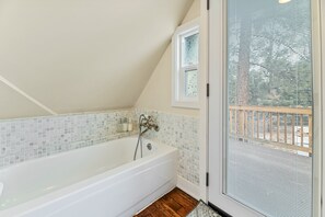 A bathroom with a white bathtub, a window with a view of a wooden deck, and a tiled backsplash.
