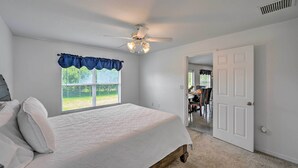 A bedroom with a bed, a ceiling fan, a window with a blue valance, and a door leading to another room. - Ranch-Style Lehigh Acres House with Big Backyard! (Lehigh Acres)